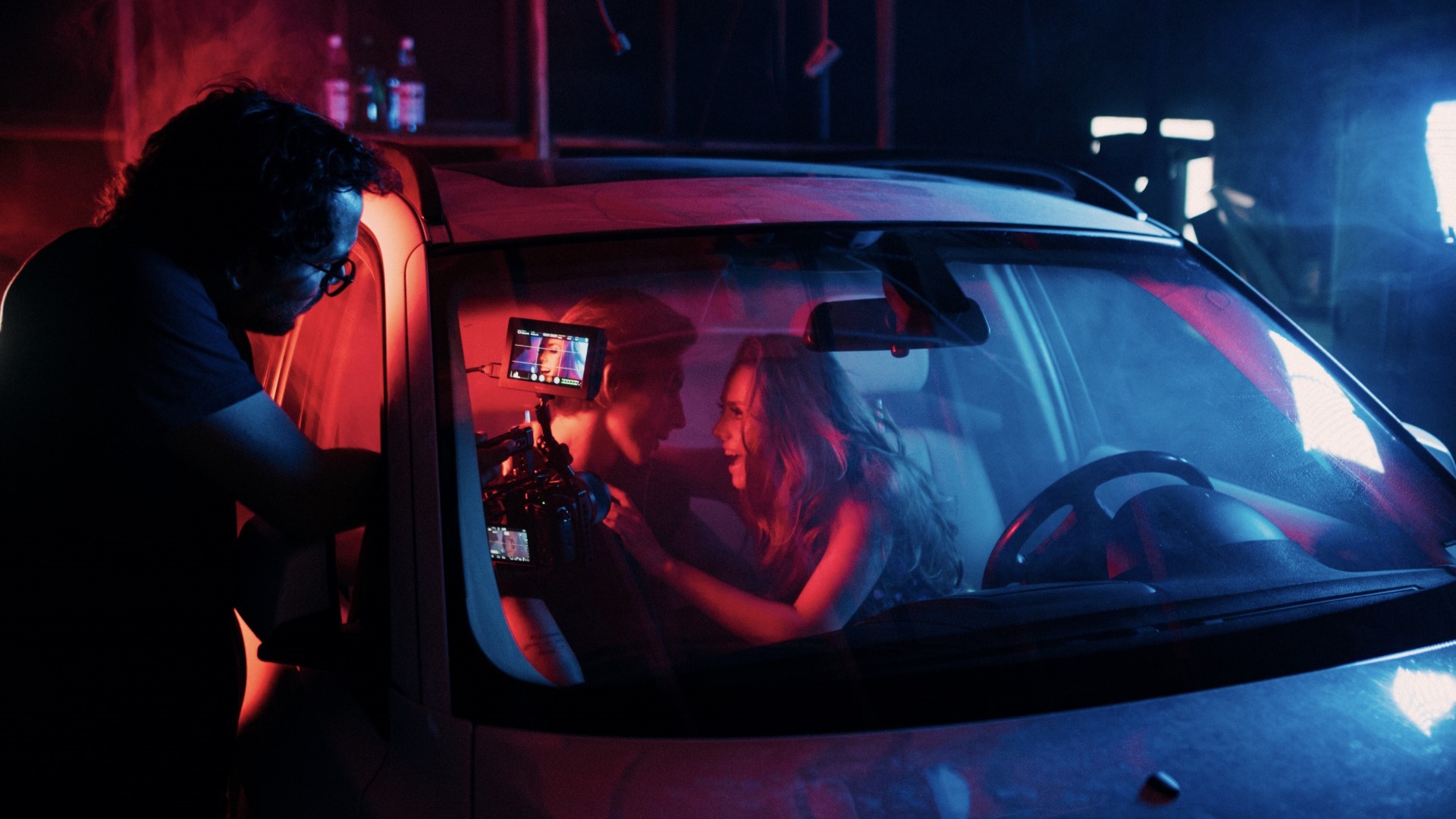 Man and Woman in a car with another man holding a camera, filming them. The car is set in a dark movie set.